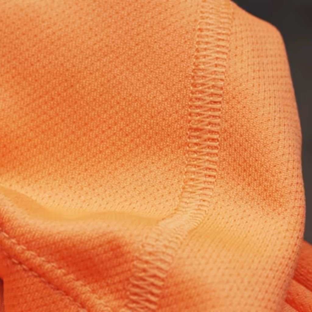 Orange athletic fabric close-up showing evenly spaced breathable perforations and visible grid-like seam stitching.
Hyper Cool Performance Fabric:
