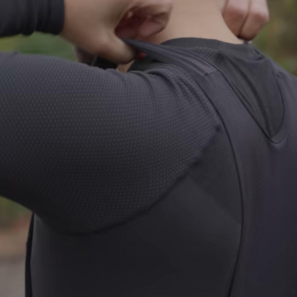 "A person's hands pulling a black long-sleeved shirt away from their neck in a rear close-up; shirt has a subtle grid-textured fabric; blurred outdoor background with muted green and brown tones."
Sweat Shield Performance Knit:
