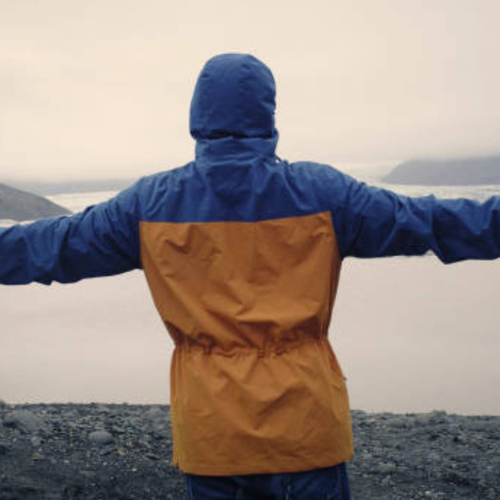 Arctic Pulse Sports Fabric: Engineered for Peak Performance 10 A person in a blue-and-mustard hooded winter jacket stands with arms outstretched on rocky terrain overlooking water and a distant shoreline under a cloudy sky.
Arctic Pulse Sports Fabric: