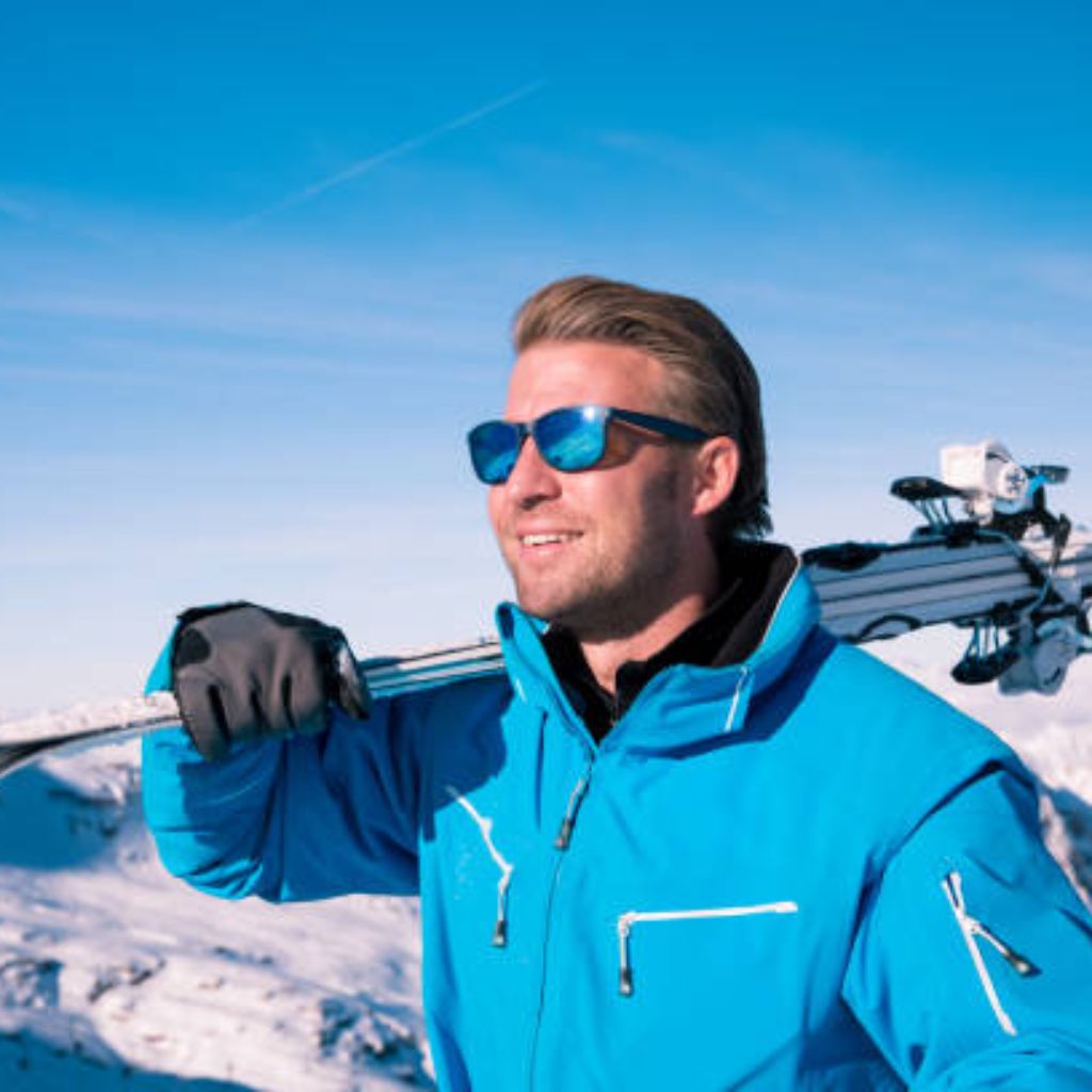 Arctic Pulse Sports Fabric: Engineered for Peak Performance 11 "A smiling man wearing a bright blue military-inspired winter jacket stands against snowy mountains and a clear blue sky. He wears reflective blue sunglasses, has skis slung over his right shoulder with visible binding hardware, and wears a grey glove on his left hand."
Arctic Pulse Sports Fabric: