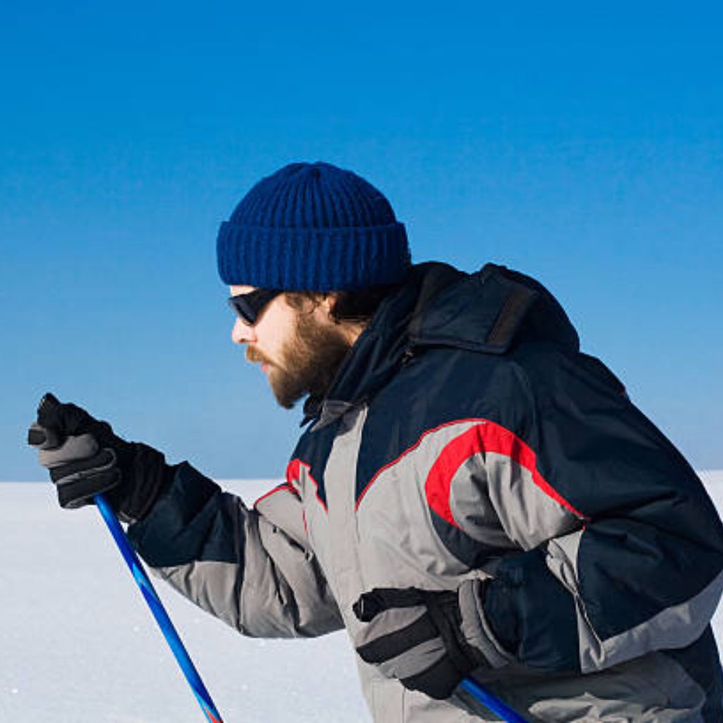 Arctic Pulse Sports Fabric: Engineered for Peak Performance 7 A person cross-country skiing on flat snow under a clear blue sky, wearing an Arctic Pulse gray-and-navy winter jacket with red accents, blue beanie, sunglasses, and black gloves.
Arctic Pulse Sports Fabric: