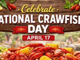 National Crawfish Day banner illustration with the headline ‘Celebrate’ in yellow script above large white text reading ‘NATIONAL CRAWFISH DAY.’ A red ribbon-shaped banner under the headline says ‘APRIL 17.’ The background is a sunny outdoor scene with people gathered by a table, while the foreground shows a heap of red crawfish, corn on the cob, lemons, and seafood on a rustic wooden surface.”
