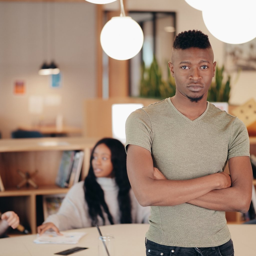 A young man stands confidently with arms crossed in a bright, cozy café while a seated woman works at a table in the background.

