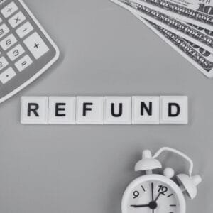 A flat lay image featuring the word "REFUND" spelled out with white tiles on a gray background. A calculator and a white alarm clock are also visible along with several one-hundred dollar bills.
