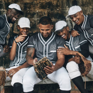 A group of five smiling baseball players in matching gray uniforms with white caps, gathered closely together on a bench. One player in the center holds a glove and looks down, while teammates lean in and laugh, showing camaraderie.