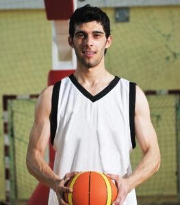 A young man standing in a gymnasium holding an orange basketball, wearing a white sleeveless basketball jersey with black trim.