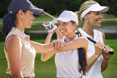 Three women on a golf course celebrate together. One woman in a beige sleeveless top and dark cap high-fives another woman in a white cap and white polo, while the third woman in a white visor smiles in the background, holding a golf club. All are on a grassy course with trees in the distance.