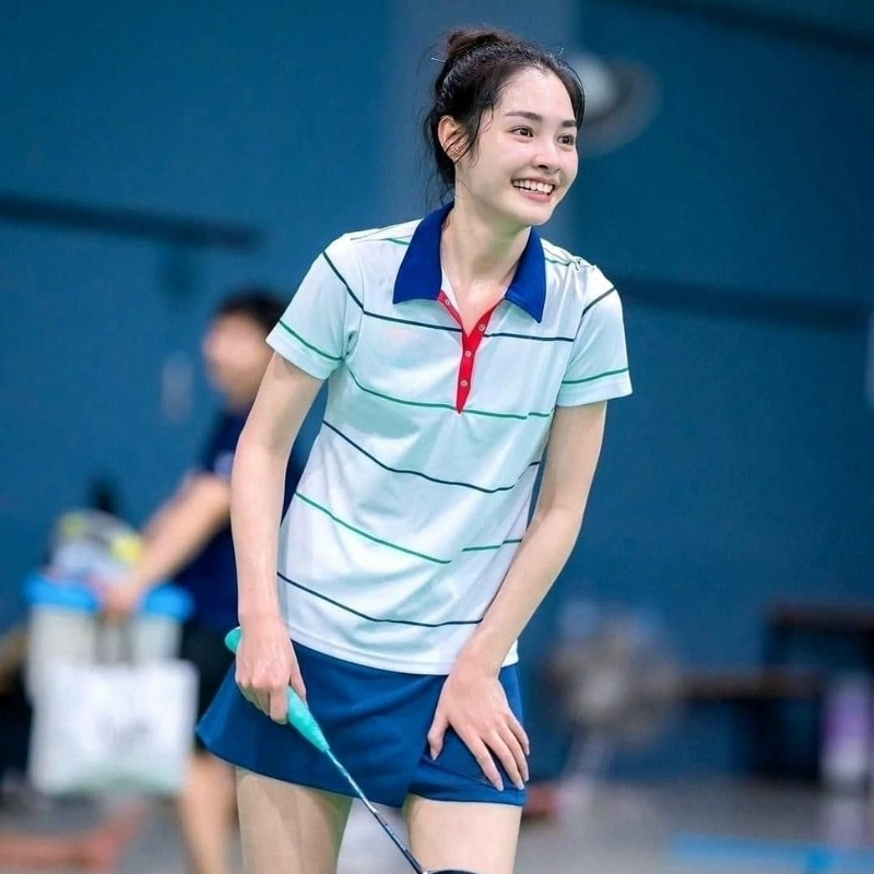 A smiling young woman wearing a light blue badminton shirt with dark blue collar and thin horizontal stripes, paired with dark blue shorts, holding a turquoise racket.