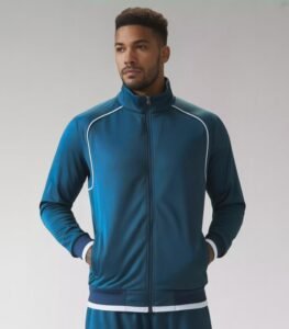 A male model wearing a blue athletic basketball jacket with a full zipper, white piping along the shoulders, and ribbed cuffs and waistband.