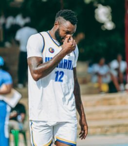 A basketball player wearing a white jersey with blue and gold trim, number 12, stands outdoors with a contemplative pose, touching his nose. He has a beard and short, curly hair, and tattoos on his arm.