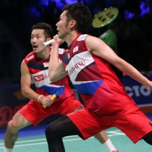 Two male badminton players in matching red, white, and blue uniforms are mid-match on an indoor court; the foreground player is preparing to strike the shuttlecock with his racquet while the teammate watches, with a blurred audience in the background.
