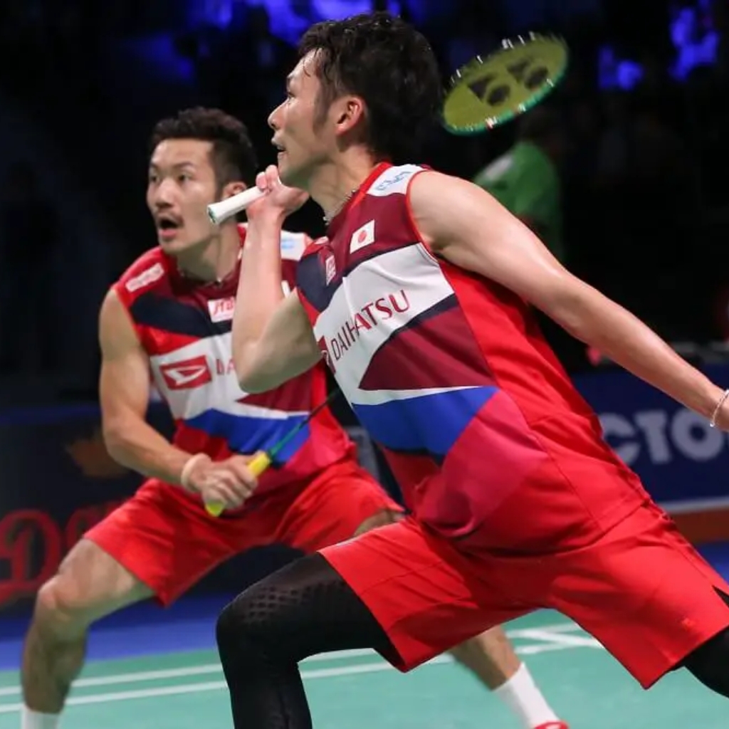 Two male badminton players in matching red, white, and blue uniforms are mid-match on an indoor court; the foreground player is preparing to strike the shuttlecock with his racquet while the teammate watches, with a blurred audience in the background.