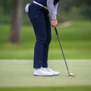 Close-up view of a golfer’s lower body on the green, wearing navy blue golf pants and white sneakers, standing at a putt with a putter in hand