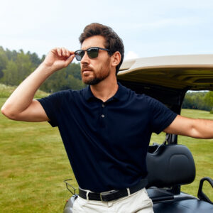  A man wearing sunglasses and a navy blue polo shirt leans against a golf cart on a sunny day. He has a beard and dark hair, and is dressed in light-colored pants. The background shows a green grassy field and trees.