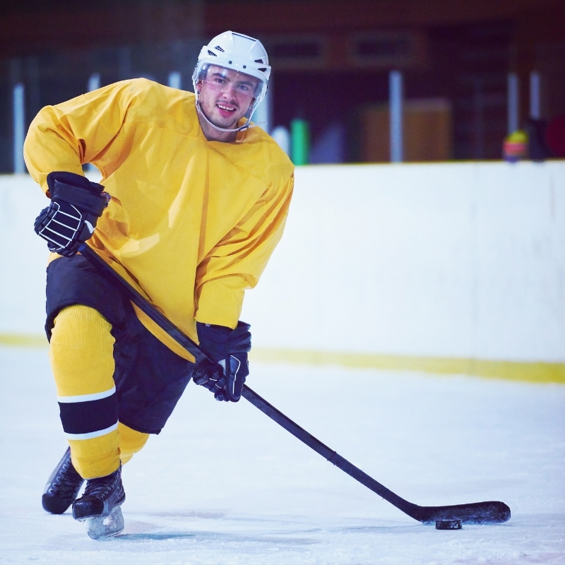 A hockey player in a bright yellow jersey and black pants glides on the ice, wearing a white helmet and black gloves, while handling a hockey stick. He smiles at the camera, with the rink boards visible in the background.