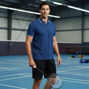 Man wearing a blue badminton polo shirt and black shorts on an indoor tennis/badminton court, holding a racket