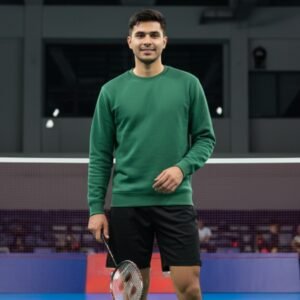 Man in a green crewneck sweatshirt holding a badminton racket indoors on a court.