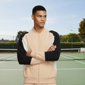 Man standing on a tennis court with arms crossed, wearing a peach-colored zip-up jacket with black and white sleeves.