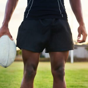 A close-up view of a rugby player from the waist down, wearing black shorts and a black shirt, holding a white rugby ball in his left hand on a grassy field.