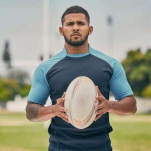  A rugby player in a two-toned blue top holds a rugby ball on a grassy field, looking focused.