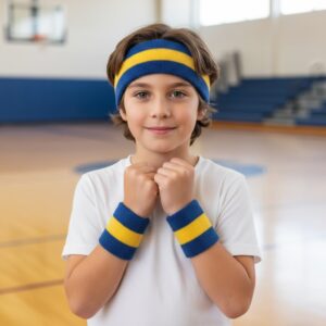 A cheerful young boy in a gymnasium wears a white T-shirt, a blue and yellow striped headband, and matching blue and yellow striped wristbands. He smiles with his fists clenched in front of him. Bleachers and a basketball court are visible in the background.