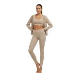 Woman standing in a light tan workout outfit consisting of a sports bra, high-waisted leggings, and a zip-up hoodie, barefoot with hands on hips