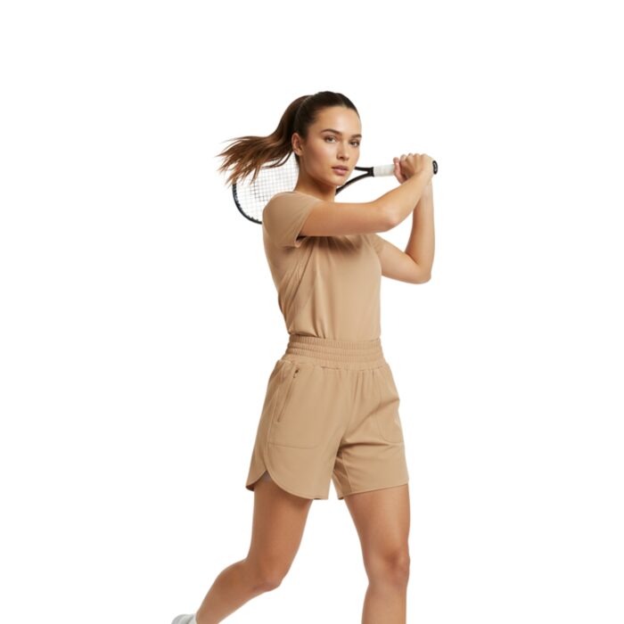 Woman wearing tan athletic shorts and matching top, mid-swing with a tennis racket on a white background. She has white socks and white tennis shoes, with her hair in a ponytail.