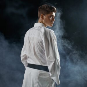 A young man wearing a white karate gi with a black belt stands with his back to the viewer, his head turned to the right, in a dark, smoky studio setting.