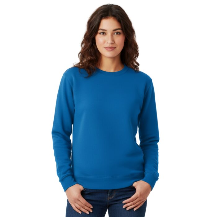 A woman stands against a plain white background, wearing a bright blue crewneck sweatshirt and dark jeans. She has her hands in her pockets