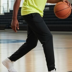 Man dribbling basketball on indoor court wearing black pants, neon yellow shirt, and white sneakers