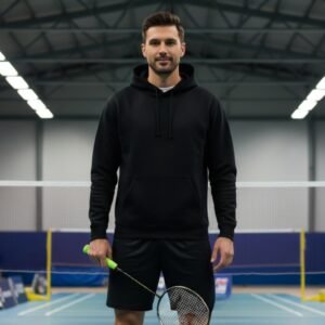Man wearing a black badminton hoodie and shorts holds a tennis-style racket in an indoor sports court.