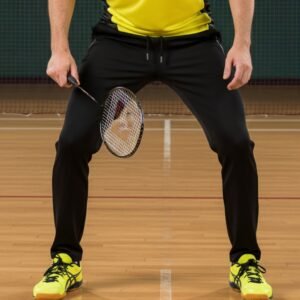 A badminton player in a yellow top and black pants holding a racket, standing on an indoor court.