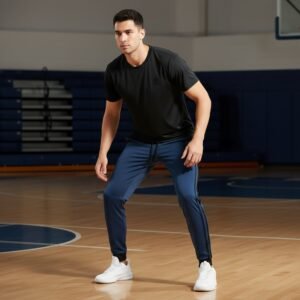 Male basketball player model wearing black basketball joggers and a sleeveless top, standing with a basketball in front of a court‑style background.