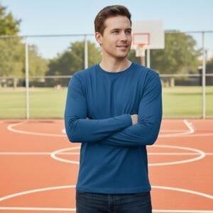 A person wearing a relaxed-fit long-sleeve crew-neck t-shirt in deep navy blue stands on a sunlit outdoor basketball court. Arms are crossed comfortably at chest height. The shirt drapes smoothly over shoulders and torso, with clean tapering at the waist. Background shows a rust-orange basketball hoop and leafy green trees in soft focus.