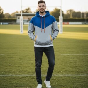 Model wearing a two-tone blue and gray pullover hoodie with orange drawstrings and yellow side pocket accents, standing on a soccer field.