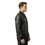 Side profile of a man wearing a black leather bomber jacket with ribbed cuffs and hem, dark wash jeans, standing against a white background.