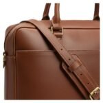 Close-up view of a brown leather briefcase showcasing the adjustable strap, stitched detailing, and gold-tone hardware.