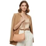 Model wearing an apricot leather shoulder bag paired with a beige blazer, satin top, and white trousers, showcasing the bag’s stylish design.