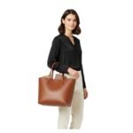 Tan leather tote bag with two top handles, worn by a model in a front carry shot