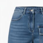 Close-up of blue denim jeans showing front pocket, button closure, stitching detail, and side pocket.