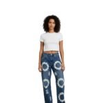 Woman wearing white crop top and blue distressed denim jeans with white floral embroidery and decorative patterns, posing against a plain white background.