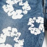 Close-up of blue denim fabric showing textured weave and hand-painted white floral patterns, with a seam visible on the right.