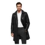 A man wearing a stylish black leather double-breasted coat over a white dress shirt and gray trousers, standing against a white background