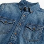 Close-up of blue denim shirt fabric showing button-down collar, buttoned placket, and two chest pockets with flaps.