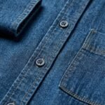 Close-up of a blue denim shirt's front placket showing two buttons, contrasting light brown stitching, and visible fabric weave texture.