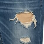 Close-up of distressed denim showing two frayed holes with loose threads and a tan fabric patch underneath.