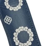 Close-up of dark blue distressed denim jeans with white floral embroidery and geometric stitch patterns, showing frayed knee tear and textured fabric.