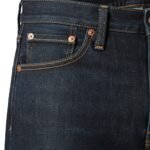 Raw denim jean close-up showing copper rivets, orange contrast stitching, and textured indigo fabric at the coin pocket seam.