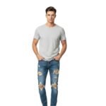 Man in gray t-shirt and ripped blue jeans standing on white background, hands in pockets.