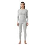 Smiling woman wearing a light grey seamless ski base layer set with long-sleeve top and leggings, standing front facing against a white background.